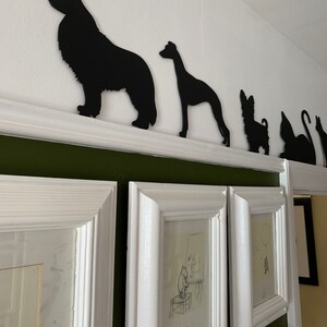 Op de afbeelding: Zwarte silhouetten van een hond, een windhond, een kleine hond en een kat gemonteerd op een witte plank tegen een groene muur. De silhouetten zijn uit zwart papier geknipt en zijn in een rij gerangschikt.