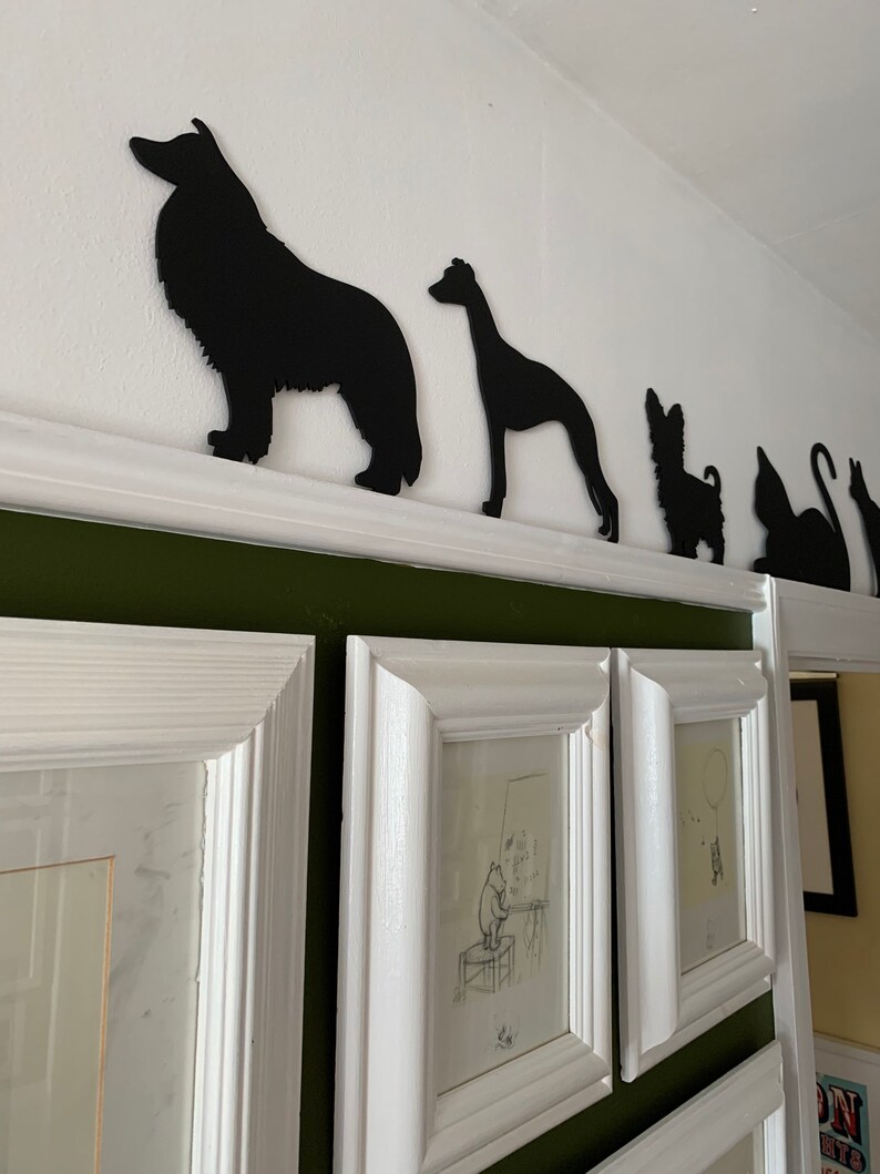 Op de afbeelding: Zwarte silhouetten van een hond, een windhond, een kleine hond en een kat gemonteerd op een witte plank tegen een groene muur. De silhouetten zijn uit zwart papier geknipt en zijn in een rij gerangschikt.