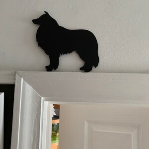 Op de afbeelding: Zwarte silhouet van een Collie-hond die op een witte deurpost staat.