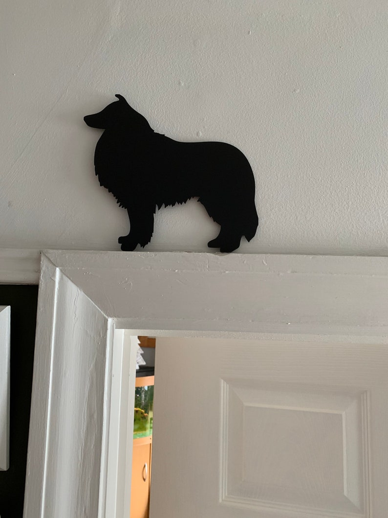 Op de afbeelding: Zwarte silhouet van een Collie-hond die op een witte deurpost staat.