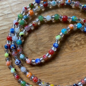 May include: Multicolored glass bead necklace with clear beads. The necklace is made of small, round beads with various patterns and colors, including red, blue, green, yellow, and white.