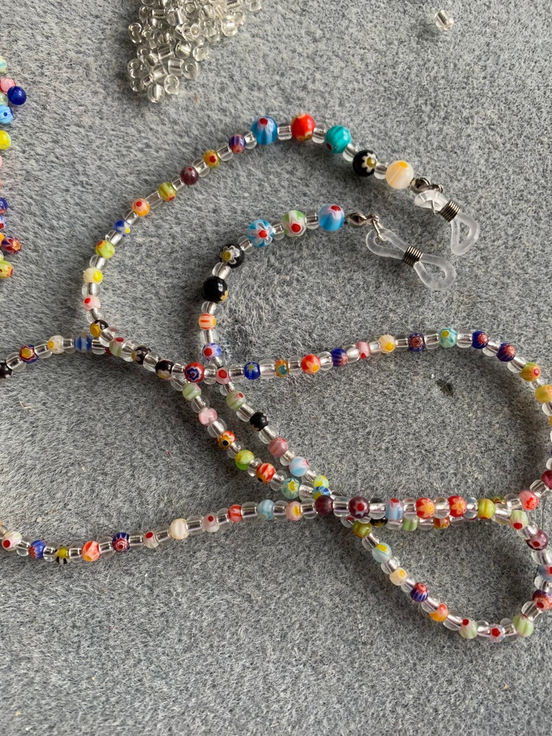 May include: A colorful beaded glasses chain with clear beads and small, colorful glass beads with floral patterns. The chain has two clear plastic clasps for attaching to glasses.