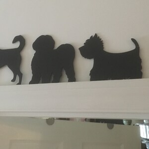 Op de afbeelding: Een rij van zes zwarte silhouetten van honden, uitgesneden op een witte muur. De silhouetten zijn van verschillende hondenrassen, waaronder een chihuahua, een terri&euml;r, een poedel, een schnauzer, een yorkie en een teckel.
