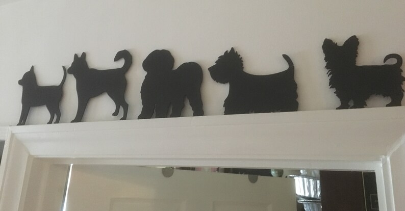 Op de afbeelding: Een rij van zes zwarte silhouetten van honden, uitgesneden op een witte muur. De silhouetten zijn van verschillende hondenrassen, waaronder een chihuahua, een terri&euml;r, een poedel, een schnauzer, een yorkie en een teckel.