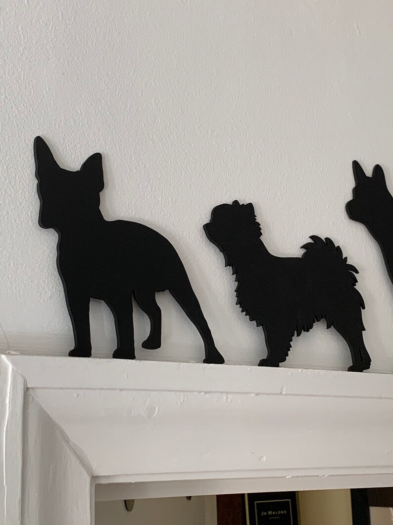Op de afbeelding: Drie zwarte silhouet-uitsnijdingen van honden op een witte muur. De honden zijn een Franse Bulldog, een Shih Tzu en een Jack Russell Terrier.