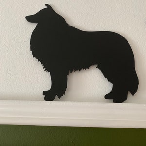 Op de afbeelding: Zwarte silhouet van een Collie-hond, uitgesneden uit hout of metaal, tegen een witte achtergrond.