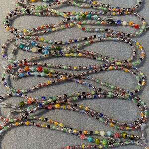 May include: A collection of colorful beaded eyeglass chains. The chains are made with small, round beads in various colors and patterns, including clear, white, black, red, green, blue, and yellow. The chains are strung on a clear cord and have metal clasps at each end.