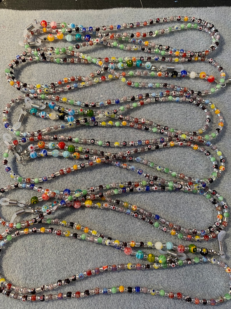 May include: A collection of colorful beaded eyeglass chains. The chains are made with small, round beads in various colors and patterns, including clear, white, black, red, green, blue, and yellow. The chains are strung on a clear cord and have metal clasps at each end.