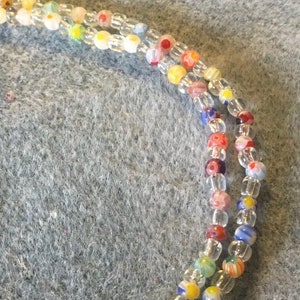 May include: A close-up of a multi-strand beaded necklace with clear, white, red, yellow, blue, and green beads. The beads have a variety of patterns, including flowers and stripes.