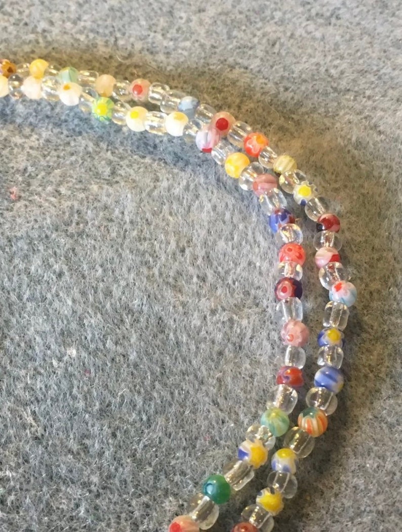 May include: A close-up of a multi-strand beaded necklace with clear, white, red, yellow, blue, and green beads. The beads have a variety of patterns, including flowers and stripes.