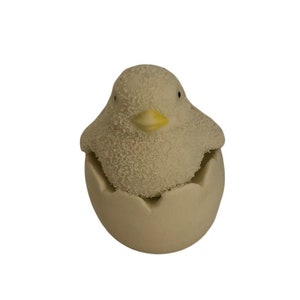 May include: A white ceramic chick hatching from a white egg. The chick has a yellow beak and black eyes.