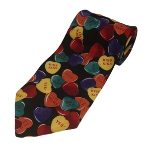 May include: A black silk tie with a pattern of colorful heart-shaped candies. The candies have various messages printed on them, including "I'm Yours", "Hug Me", "Kiss Kiss", and "Yes!"