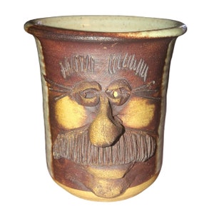 Könnte beinhalten: Eine braune und gelbe Keramiktasse mit einem auf der Vorderseite modellierten Gesicht. Das Gesicht hat eine große Nase, buschige Augenbrauen und einen Schnurrbart.