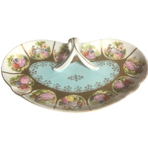 May include: A white porcelain serving tray with a blue and gold design. The tray has a handle and features a repeating pattern of couples in a garden setting.