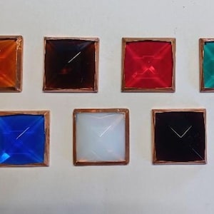 May include: Seven square-shaped, colorful glass pieces with copper frames. The colors include orange, dark brown, red, teal, blue, white, and black. Each piece has a pyramidal shape and is set against a white background.