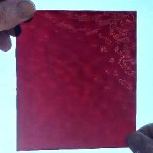 May include: A square piece of ruby red stained glass is held up against a light blue background. The glass has a textured surface and is semi-transparent, allowing light to pass through. The edges are clean and straight.