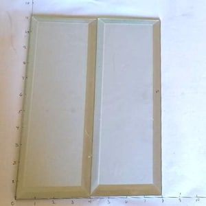 May include: Two rectangular, off-white trays with a raised border are placed side by side. The trays are approximately 10 inches long and 5 inches wide. The background is a white surface with a ruler drawn on it.
