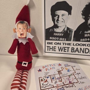 May include: A red and white elf doll with a cutout of a surprised face. The doll is next to a "Wet Bandits" poster and a "Battle Plan" drawing with text and illustrations. The elf has striped legs and a black belt.