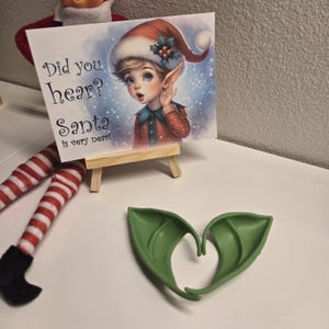 May include: A Christmas elf with red and white striped leggings and black shoes. The elf is holding a sign that says "Did you hear? Santa is very near!" The sign is on a small wooden easel. There are also two green elf ears on the table.