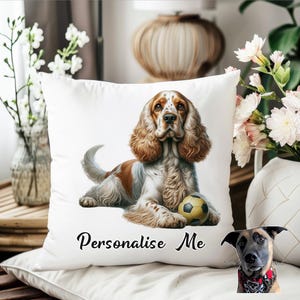 Può includere: Cuscino quadrato bianco con un'illustrazione di Cocker Spaniel. Il cane è marrone, bianco e marrone chiaro, con un pallone da calcio. Il testo "Personalise Me" è stampato in corsivo nero. Il cuscino è su una superficie bianca.