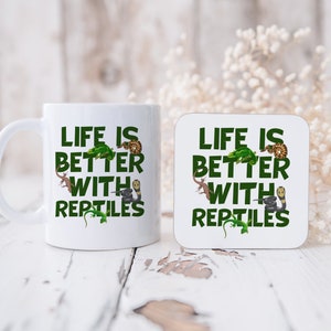 Può includere: Set di tazza e sottobicchiere in ceramica bianca con testo verde che recita "La vita è migliore con i rettili". Il testo è circondato da immagini di vari rettili, tra cui un serpente, una lucertola e una tartaruga.