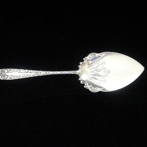 May include: A silver cake server with an ornate handle and a wide, leaf-shaped blade. The handle features a detailed, swirling pattern, and the blade has a decorative edge. The server is designed for serving cakes and pastries.