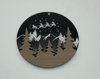 Puede incluir: Un adorno redondo de madera negro y marrón con una silueta blanca de una escena de bosque de invierno con un trineo y renos volando por el aire. El adorno tiene un agujero en la parte superior para colgar.