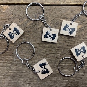 May include: Six wooden keychains with different hand signs for the letters I, L, O, and Y in American Sign Language. Each keychain has a silver key ring.