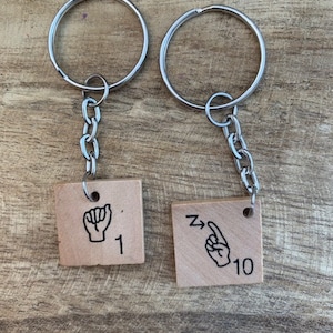 May include: Two silver keychains with wooden tags. The tags are square and have black illustrations of hands signing the numbers 1 and 10. The number 1 tag has the text "1" and the number 10 tag has the text "10".