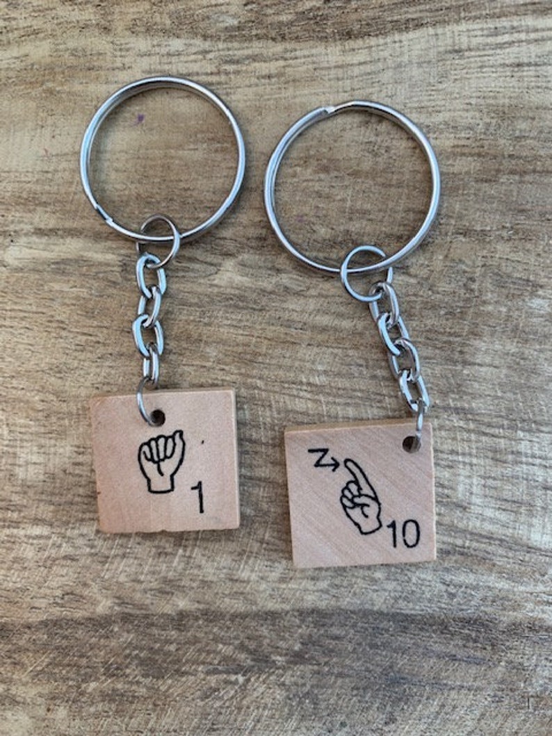 May include: Two silver keychains with wooden tags. The tags are square and have black illustrations of hands signing the numbers 1 and 10. The number 1 tag has the text "1" and the number 10 tag has the text "10".