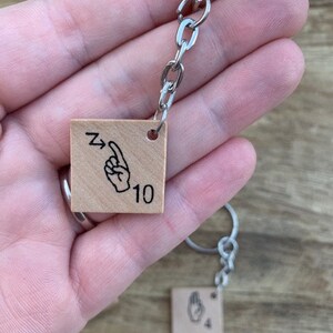 May include: A wooden keychain with two square tags. The tags feature a hand sign for the number 10 and the number 4. The keychain is attached to a silver chain.