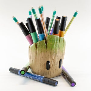 Puede incluir: Un porta lápices con forma de Groot de color verde y marrón con una variedad de marcadores y bolígrafos de colores que sobresalen de la parte superior.