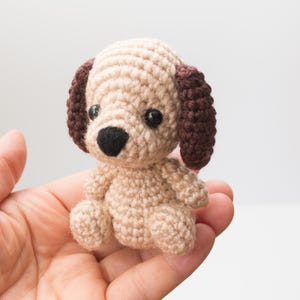 Puede incluir: Figura de perro de ganchillo en beige y marrón. El perro tiene orejas marrones oscuras, una nariz negra y ojos negros. El juguete hecho a mano está sentado en la mano de una persona.