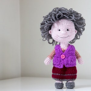May include: A crocheted doll with gray hair, a pink face, a purple vest with a pink button, a burgundy skirt with gray stripes, and gray shoes.