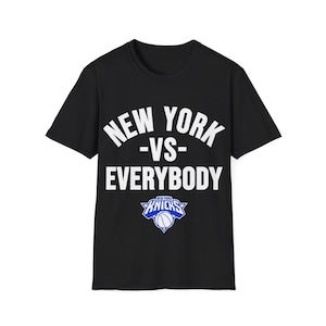May include: A black short-sleeved t-shirt featuring the white text NEW YORK -VS- EVERYBODY printed in a bold, arched font. Below the text is a blue and white New York Knicks basketball team logo, which includes a basketball graphic.
