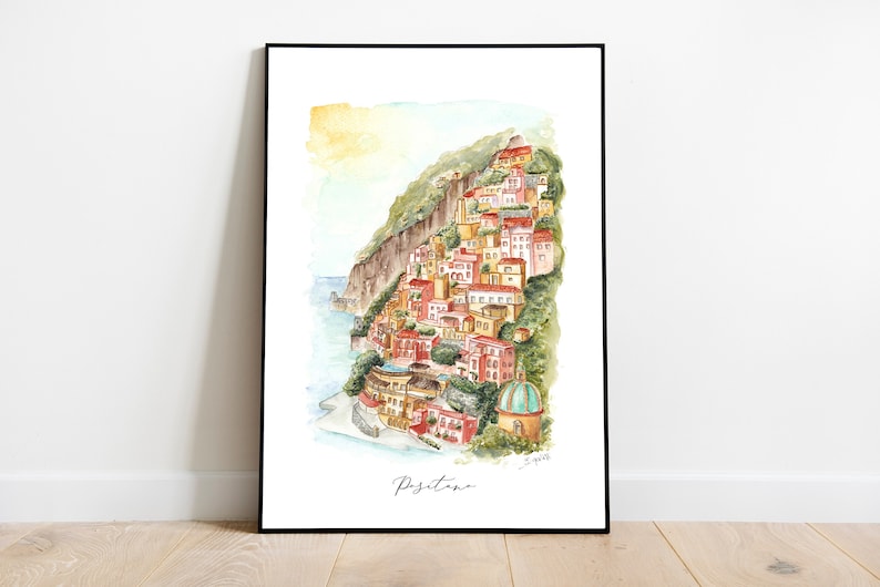 May include: Watercolor painting of Positano, Italy, featuring colorful buildings cascading down a hillside towards the sea. The artwork is framed in black and includes the town's name in cursive script.