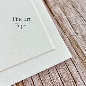 May include: Close-up of two sheets of fine art paper, slightly overlapping. The paper is a soft, off-white color. The words "Fine art Paper" are printed in a simple, elegant font on the top sheet. The background is a wooden surface.