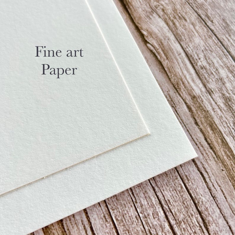 May include: Close-up of two sheets of fine art paper, slightly overlapping. The paper is a soft, off-white color. The words "Fine art Paper" are printed in a simple, elegant font on the top sheet. The background is a wooden surface.