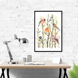 May include: A framed watercolor print featuring several colorful birds perched on branches. The birds include a red bullfinch, a blue tit, a robin, and a goldfinch. The artwork is displayed on a wooden desk with a silver lamp and a white brick wall background.
