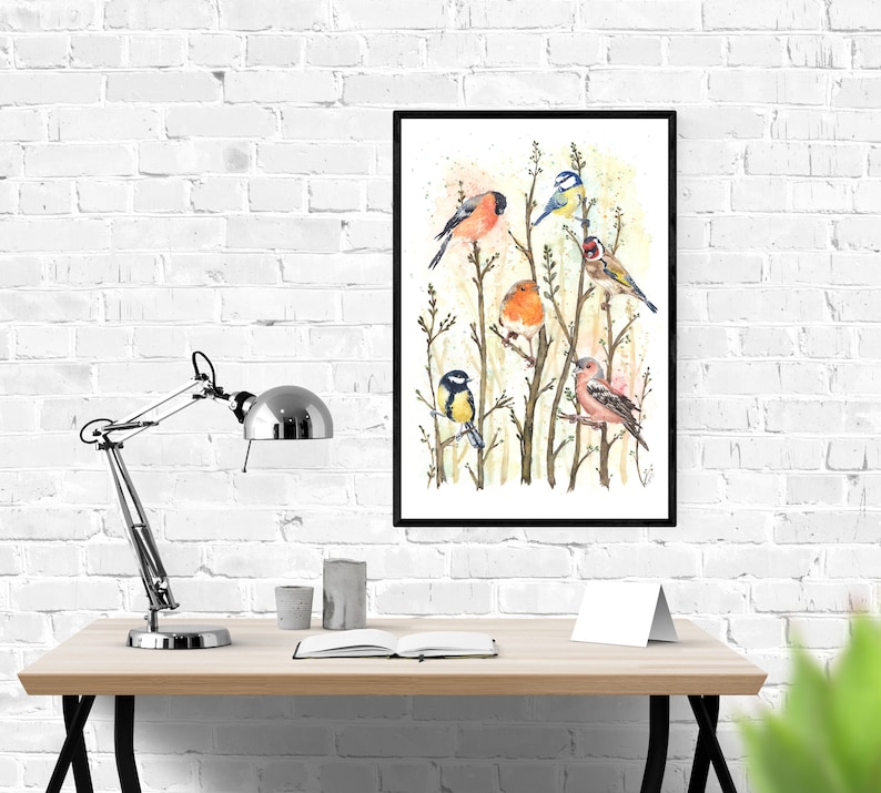 May include: A framed watercolor print featuring several colorful birds perched on branches. The birds include a red bullfinch, a blue tit, a robin, and a goldfinch. The artwork is displayed on a wooden desk with a silver lamp and a white brick wall background.