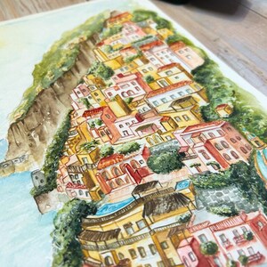 以下が含まれることがあります： 丘を下るカラフルな建物が特徴の、海岸沿いの町の水彩画。このアートワークは、黄色、オレンジ、赤などのさまざまな暖色系と、緑の葉と青い水が特徴です。絵は白い表面にあります。