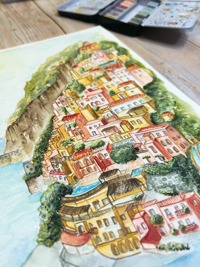 以下が含まれることがあります： 丘を下るカラフルな建物が特徴の、海岸沿いの町の水彩画。このアートワークは、黄色、オレンジ、赤などのさまざまな暖色系と、緑の葉と青い水が特徴です。絵は白い表面にあります。