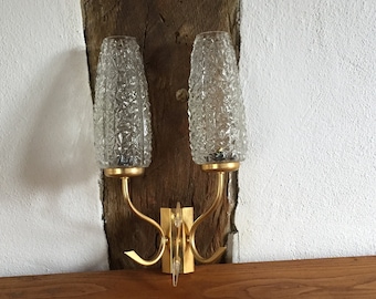 Può includere: Un'applique da parete dorata con due paralumi in vetro trasparente con un motivo testurizzato. L'applique è montata su una parete in legno.