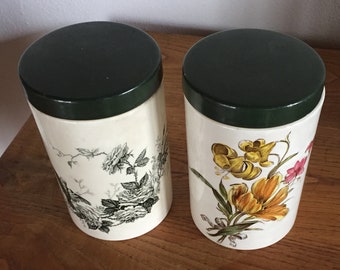 Può includere: Due barattoli in ceramica con coperchi verde scuro. Uno presenta un motivo floreale in bianco e nero, l'altro una composizione floreale colorata. I barattoli sono cilindrici e poggiano su una superficie di legno.