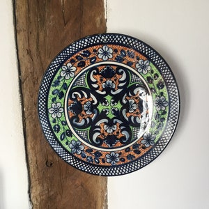 Può includere: Un piatto in ceramica decorativo con un motivo floreale blu, verde, arancione e bianco. Il piatto è appeso a una parete bianca.