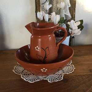 Peut inclure: Un ensemble de pichet et de bol en terre cuite avec des motifs floraux blancs. Le pichet a une anse et un bec verseur. Le bol est rond et a une légère lèvre. L'ensemble est posé sur une dentelle blanche crochetée.