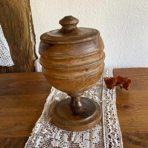 May include: A brown wooden lidded container with a rounded, ribbed body and a pedestal base. The object features a smooth, polished finish with visible wood grain patterns and a small, circular knob handle centred on the top of the lid.