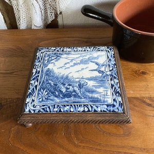 Puede incluir: Un azulejo de cerámica azul y blanco con una escena de caza, enmarcado en madera con un borde tallado. El azulejo está sobre una superficie de madera.