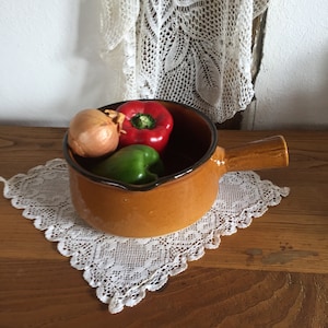 Peut inclure: Un pot en céramique brun avec une poignée, rempli d'un poivron rouge, d'un poivron vert et d'un petit oignon. Le pot est posé sur une dentelle blanche en crochet sur une surface en bois.