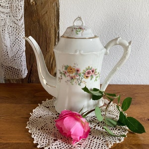 Op de afbeelding: Witte porseleinen theepot met een bloemenpatroon en gouden rand. De theepot heeft een gebogen handvat en een tuit. Een roze roos ligt op een gehaakt onderzetter.